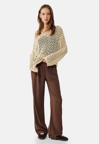 Maglione cremoso lavorato a maglia con una trama larga e maniche ampie, abbinato a pantaloni larghi marroni e indossato con delle scarpe slip-on scure.