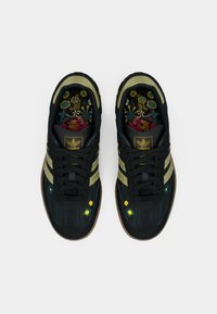Μαύρο αθλητικό παπούτσι με πράσινες και χρυσές ρίγες, υφασμάτινο επάνω μέρος με υφή και πάτος με λουλουδένιο σχέδιο. Διαθέτει κορδόνια και λαστιχένια σόλα.