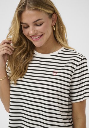 Mladá žena s dlhými blond vlasmi sa usmieva, má na sebe čierno-biele pruhované tričko s malým červeným srdcom na hrudi a zlaté kruhové náušnice.