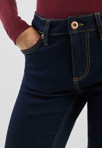 Tmavomodré riflové nohavice s viditeľným prešívaním, medeným gombíkom a prednými vreckami. Pás je mierne zvýšený a priliehavý.