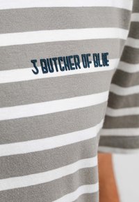 Grau-weiß gestreiftes Kurzarmhemd mit weicher Textur; verfügt über eine blaue Stickerei mit der Aufschrift "J BUTCHER OF BLUE" auf der Brust.