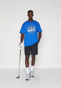 Vyras nešiojantis mėlyną Nike golfą marškinėlį, juodas šortus, baltą golfą pirštinę ir sportbačius, stovi su golfu lazdos paprastoje baltos spalvos erdvėje.
