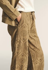 Pantalons khaki avec un motif texturé de hexagones et des découpes florales. Taille élastique avec cordon de serrage, associés à un haut vert olive.