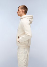 Felpa beige con cordini, tasca a marsupio e orlo a coste. Abbinata a pantaloni cargo beige chiaro con tasche laterali. Vista di profilo.