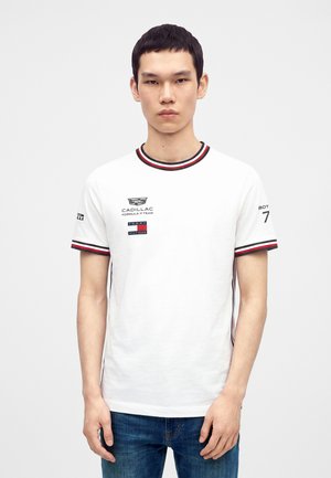 Νεαρός άνδρας φοράει λευκό μπλουζάκι Tommy Hilfiger με λογότυπα της ομάδας Cadillac Formula 1 και μπλε τζιν, στέκεται μπροστά σε ένα μονόχρωμο φόντο.
