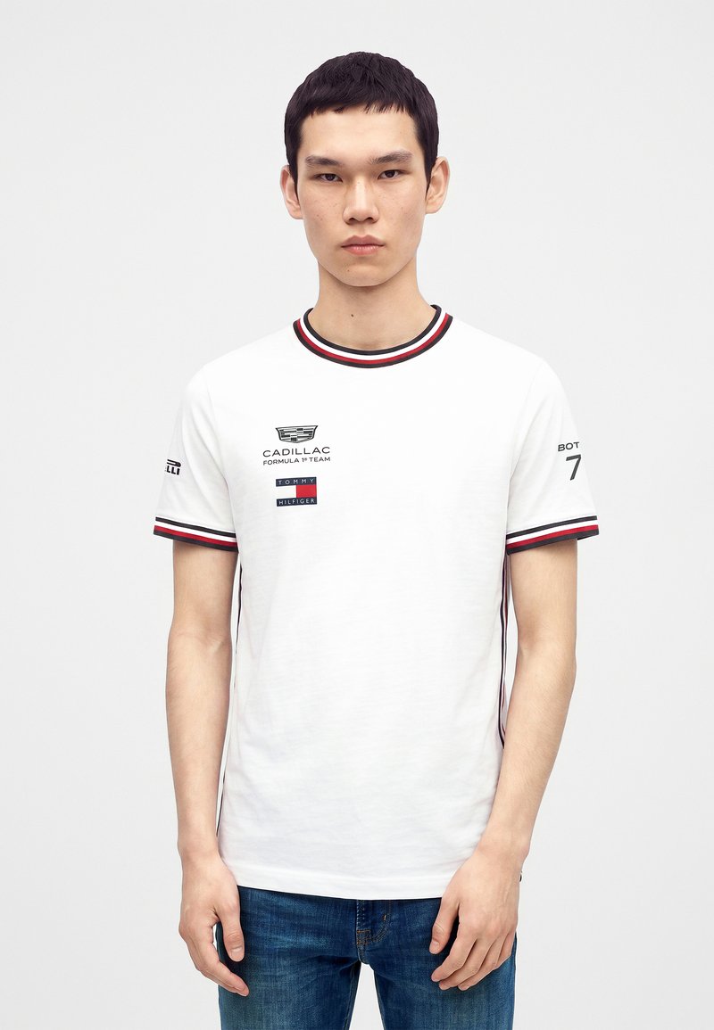 Νεαρός άνδρας φοράει λευκό μπλουζάκι Tommy Hilfiger με λογότυπα της ομάδας Cadillac Formula 1 και μπλε τζιν, στέκεται μπροστά σε ένα μονόχρωμο φόντο.