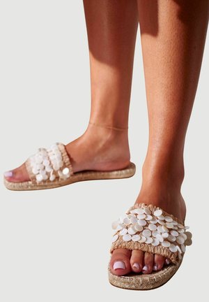 Beige Slipper-Sandalen mit großen weißen Muschel-Akzenten, einem gewebten Obermaterial und einer strukturierten Jutesohle. Offenes Zehendesign mit weichem Stoff.