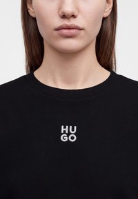 Жена, носеща черна блуза с кръгло деколте и малък бял текст "HUGO", бродиран в центъра на гърдите.