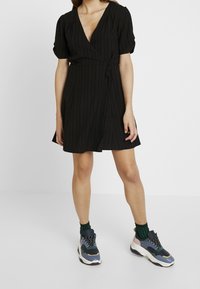 Schwarzes Wickelkleid mit vertikalen Streifen, kurzen Ärmeln und einem Bindeband in der Taille, kombiniert mit mehrfarbigen Turnschuhen und grünen gemusterten Socken.