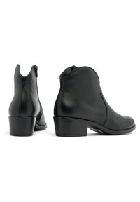 Un par de botas de cuero negro hasta el tobillo con tacones bajos, una mostrando la vista lateral y la otra trasera con una cremallera visible sobre un fondo blanco.