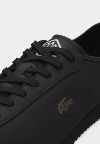 Fekete bőrből készült sportcipő fekete fűzőkkel, texturált talppal és egy kis zöld hímzett krokodil logóval az oldalán.