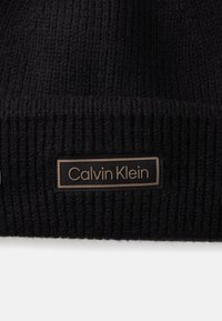 Musterniga mustriga müts, millel on silmapaistev Calvin Kleini logo silt ristkülikukujulises disainis, millel on kontrastne äär.