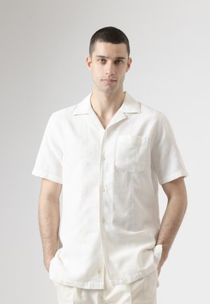 Joven con cabello corto y oscuro, vestido con una camisa blanca de manga corta con bolsillo en el pecho, de pie con las manos en los bolsillos, fondo neutro.