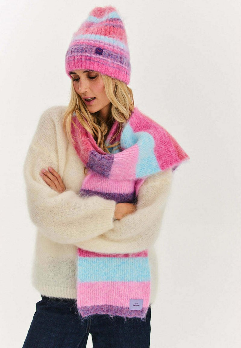 Zestaw dzianinowej czapki i szalika w paski w odcieniach różu, fioletu i niebieskiego. Miękka, puszysta faktura; szalik ozdobiony jest pasami kolorów i małą metką z logo.