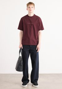 Maroon cotton t-shirt with embroidered logo, paired with dark blue wide-leg jeans and black sneakers. Model holds a black tote bag.