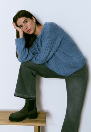 Suéter de punto azul claro con textura acanalada, combinado con jeans grises con dobladillos deshilachados y botas de ante negras hasta el tobillo sobre un taburete de madera.