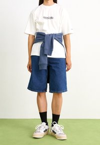 Camiseta blanca de manga corta con el logo "Columbia", combinada con pantalones cortos de mezclilla azul hasta la rodilla y un sudadera azul atada a la cintura.