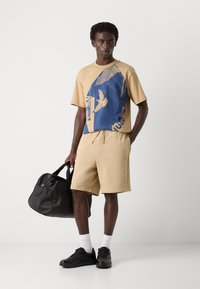 T-shirt en coton beige avec un design graphique bleu, associé à un short beige. Sac de sport noir et baskets noires avec des chaussettes blanches.