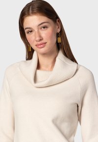 Pull à col cheminée de couleur crème, en tissu tricoté doux, avec un col côtelé et une texture lisse, présentant une silhouette ajustée.