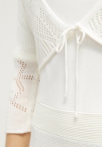 Vit stickad topp med spetsdesign, med knytning, ribbad struktur vid midjan och detaljerade mönster på ärmarna.