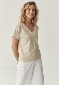 Top beige de cuello en V con mangas cortas, que presenta un detalle de rayas texturizadas en el escote y el dobladillo, combinado con pantalones blancos de corte ajustado.