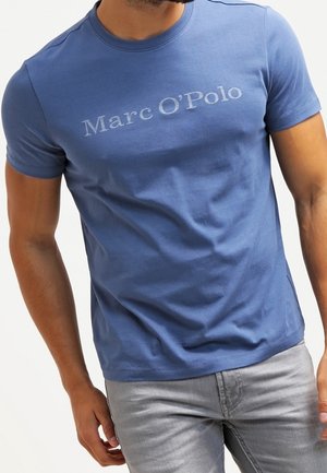 Mand iført en tætsiddende blå Marc O'Polo t-shirt og lysegrå jeans, stående med den ene hånd afslappet ned langs siden.