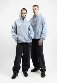 Šviesiai mėlynos gobtuvinės striukės su priekinėmis kišenėmis ir logotipu, derinamos su juodais plačiais džinsais ir juodomis avalynėmis. Lygus audinys, laisvas siluetas ir elastiniai rankogaliai.