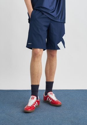 Άτομο που φοράει ναυτικό μπλε σορτς με λευκό λογότυπο, μαύρες κάλτσες και κόκκινα αθλητικά παπούτσια Adidas, όρθιο σε μπλε μοκέτα.