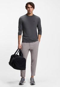 Langärmeliges graues Hemd mit dezentem Logo, kombiniert mit hellgrauen Hosen und grauen Sportschuhen, hält eine schwarze Reisetasche.