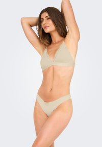 Vrouw poseert in een beige bralette met kant en bijpassende hoge slipjes tegen een effen lichte achtergrond.