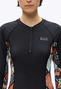 Kobieta ubrana w czarny top do pianki Billabong z przednim zamkiem błyskawicznym oraz kolorowymi, wzorzystymi panelami bocznymi i rękawami.