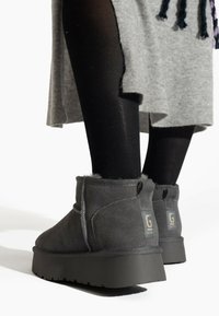 Botas de ante gris a la altura del tobillo con forro de felpa, suelas de goma planas y un detalle de logo en el talón, combinadas con leggings negros y un vestido gris.