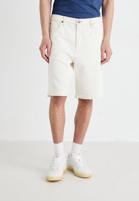 Shorts en denim blanc avec une coupe décontractée, dotés d'une fermeture à bouton et à zip, ainsi que d'un design classique à cinq poches. Associés à des baskets blanches.
