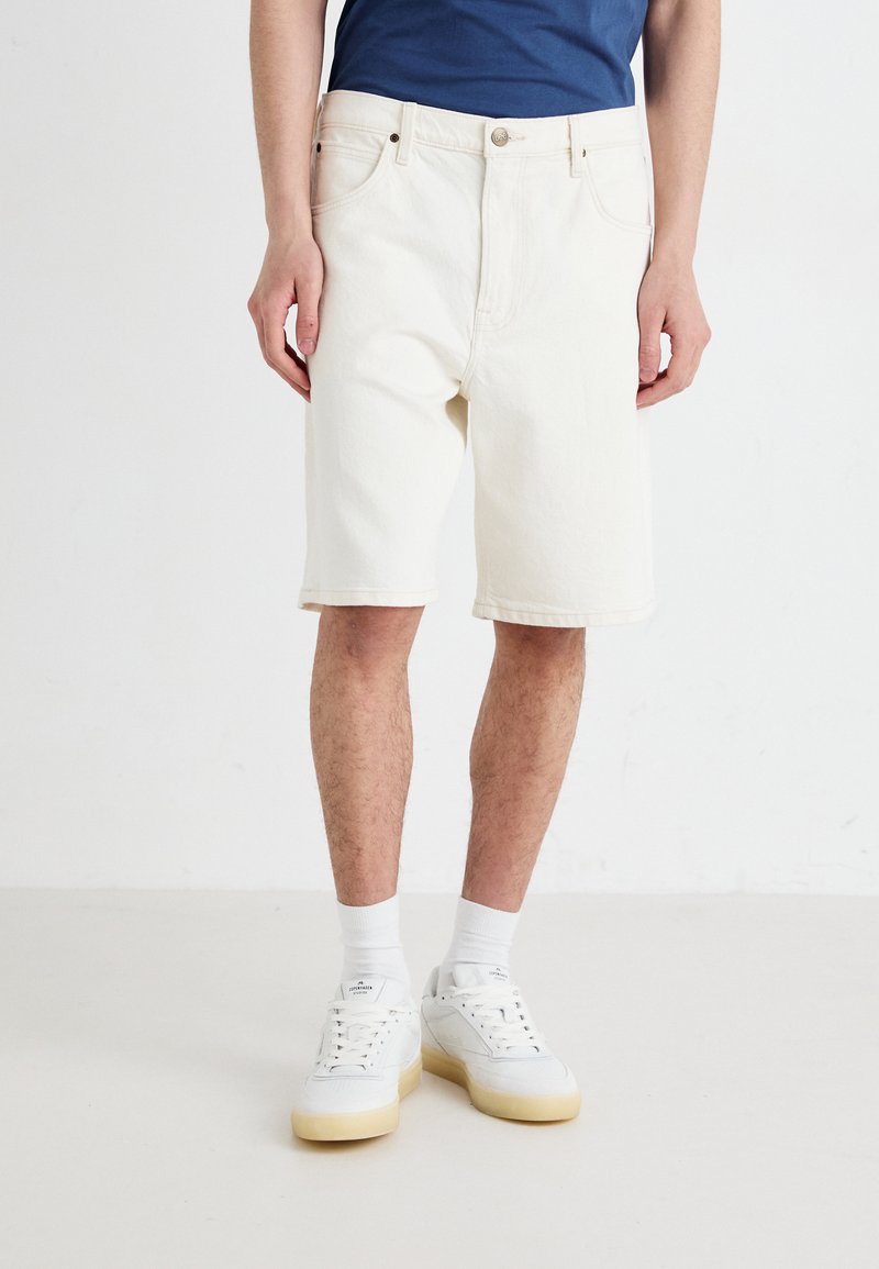 Shorts en denim blanc avec une coupe décontractée, dotés d'une fermeture à bouton et à zip, ainsi que d'un design classique à cinq poches. Associés à des baskets blanches.
