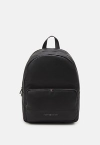 Mochila negra de Tommy Hilfiger con bolsillo frontal con cremallera y asa superior, mostrada sobre un fondo blanco.