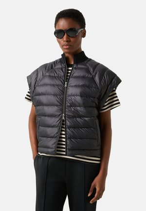 Person iført sorte solbriller, en sort vatteret vest over en sort-hvidstribet skjorte og sorte bukser mod en ensfarvet baggrund.