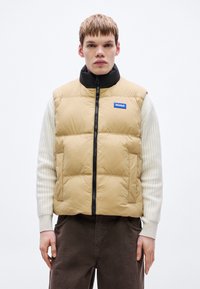 Beež joped vest musta kõrge kraega, ees tõmblukk ja logomärgiga plaastriga. Kantud tekstuurse valge pikkade varrukatega särgi ja pruunide pükstega.