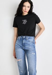 Juodas medvilnės marškinėlis su baltu "Calvin Klein Jeans NYC" logotipu. Suderinta su šviesiai mėlynomis nublukusiomis džinsinėmis kelnėmis, turinčiomis suplėšytas detales.
