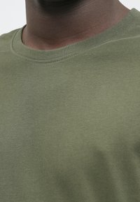 Tuvumā redzams cilvēks, kurš valkā olīvzaļo krekla ar apaļo kakla izgriezumu, koncentrējoties uz kakla aploci un augšējo krūšu daļu.