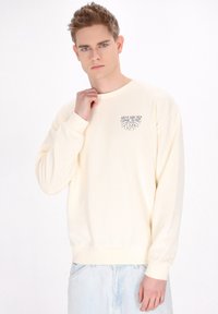 Lichtbeige sweatshirt met lange mouwen en ronde hals, voorzien van een klein zwart grafisch ontwerp op de borst, zachte textuur en geribde manchetten.