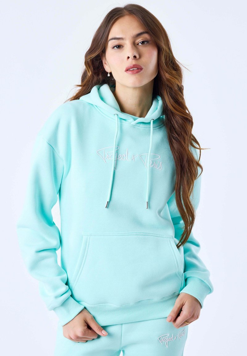 Jeune femme aux longs cheveux bruns portant un sweat à capuche bleu clair et un pantalon assorti avec un texte blanc brodé.