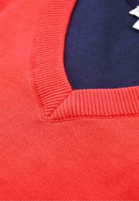 Nahaufnahme eines korallenroten V-Ausschnitt-Pullovers mit geripptem Ausschnitt und strukturiertem Strickstoff über einem dunklen Innenkleidungsstück.