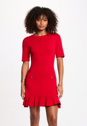 Kvinde med krøllet hår iført en rød kjole med korte ærmer, flæsekant og dekorative knapper på lommen, stående mod en hvid baggrund.