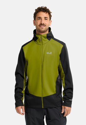 Mann trägt eine grün-schwarze Jack Wolfskin Kapuzenjacke mit Frontreißverschluss und Reißverschlusstaschen an den Seiten, steht vor einem einfarbigen Hintergrund.
