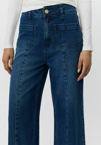 Široke tamnoplave traperice visokog struka s prednjim džepovima i kontrastnim šavovima. Teksturirana tkanina, ojačani šavovi i detalji s zlatnim dugmetom.