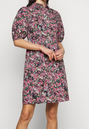 Mujer con un vestido corto floral con mangas abullonadas y cuello, con las manos ligeramente entrelazadas delante, de pie frente a un fondo liso.