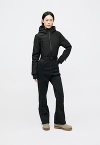 Mulher em pé com a mão na anca, vestindo uma jaqueta preta com capuz, calças pretas com cinto e botas bege, em fundo branco.