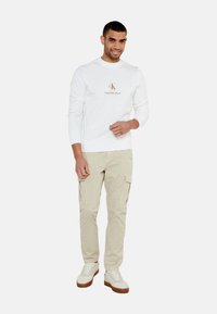 Sudadera blanca de cuello redondo con logo frontal, pantalones cargo beige y zapatillas beige. Ajuste casual, material de algodón, detalles de costura visibles.