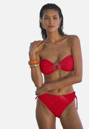 Femeie cu părul ud, purtând un costum de baie roșu fără bretele, cu un detaliu metalic în formă de soare, pozând cu brățări colorate pe brațul stâng, pe un fundal simplu.