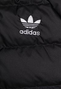 Svart puffjacka med tyg som visar den vita broderade Adidas-logotypen med tre bladliknande ränder ovanför varumärket. Slät textur.